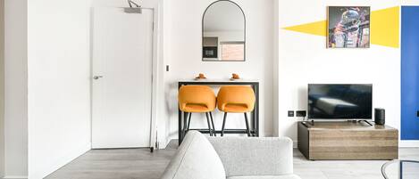 A modern living room with a sofa, a dining area with two chairs, a TV, and a wall-mounted mirror.