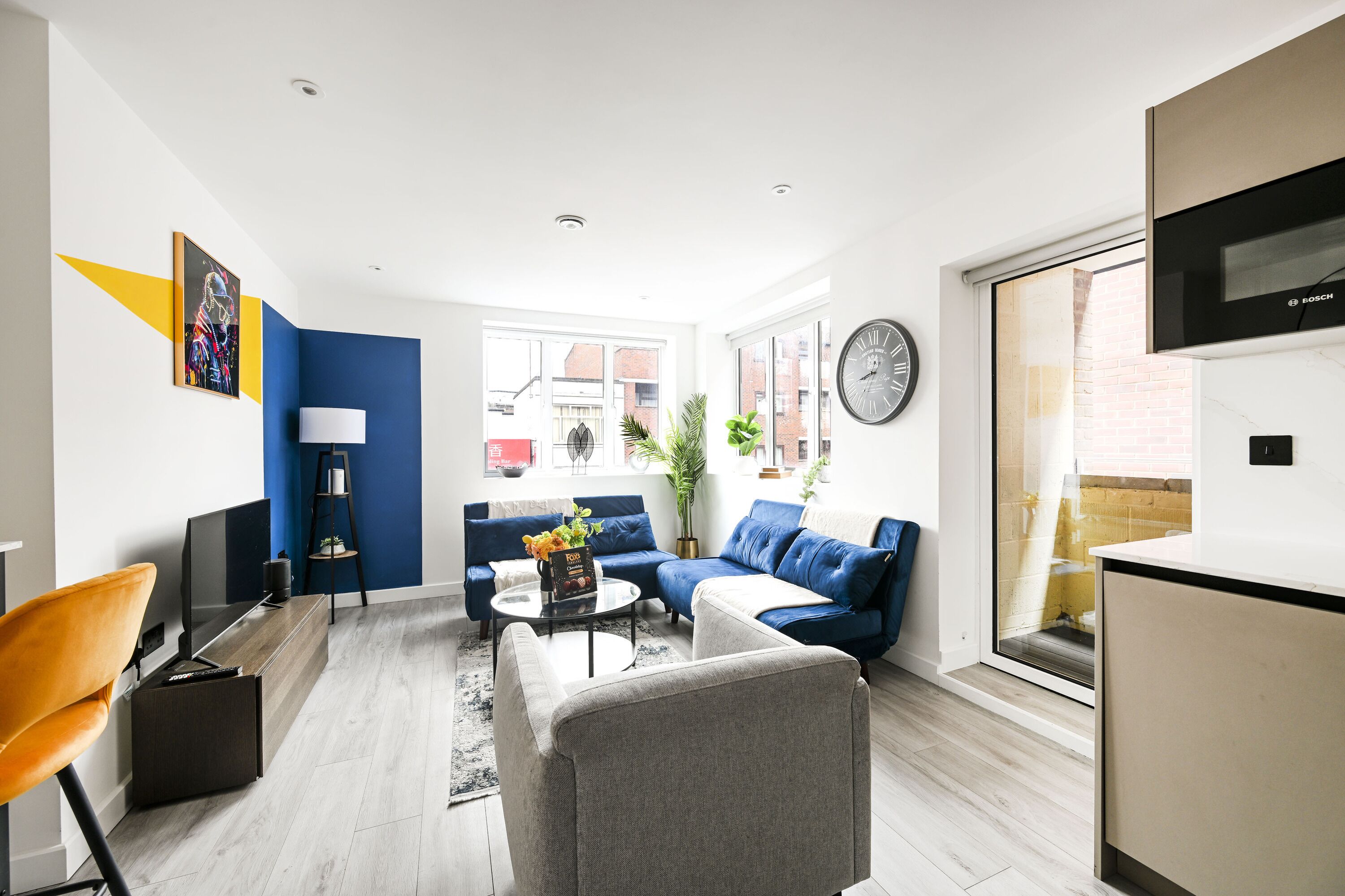 A modern living room with a sofa, armchair, and coffee table. There is a television, a clock, and a window with a view of buildings.