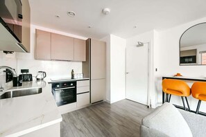 A modern kitchen with a built-in oven, a sink, and a coffee maker. There are two orange chairs and a white sofa in the living area. - Sleeps 6 | Hendon | Parking + Fast WiFi (London)