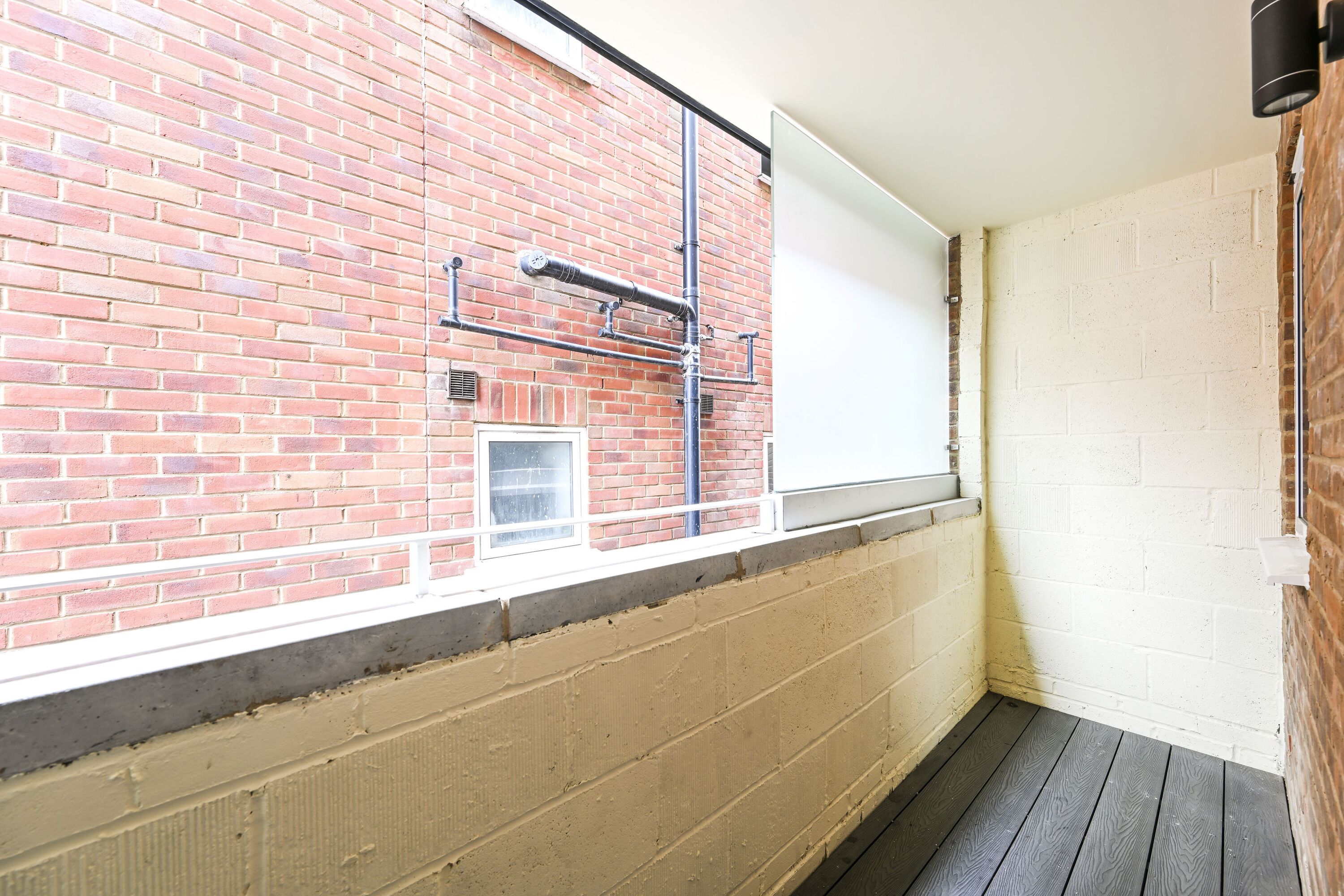 A room with a brick wall, a window with a white blind, and a wooden floor.