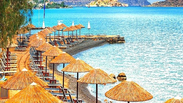 A beach with rows of sun loungers and thatched umbrellas, a pier extending into the water, and a mountainous backdrop. - BH1041 - A - Summer Home (Agios Nikolaos)
