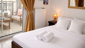 A hotel room with a bed, two pillows, a nightstand with a lamp, a window with curtains, a chair, and a radiator. - BH1041 - A - Summer Home (Agios Nikolaos)