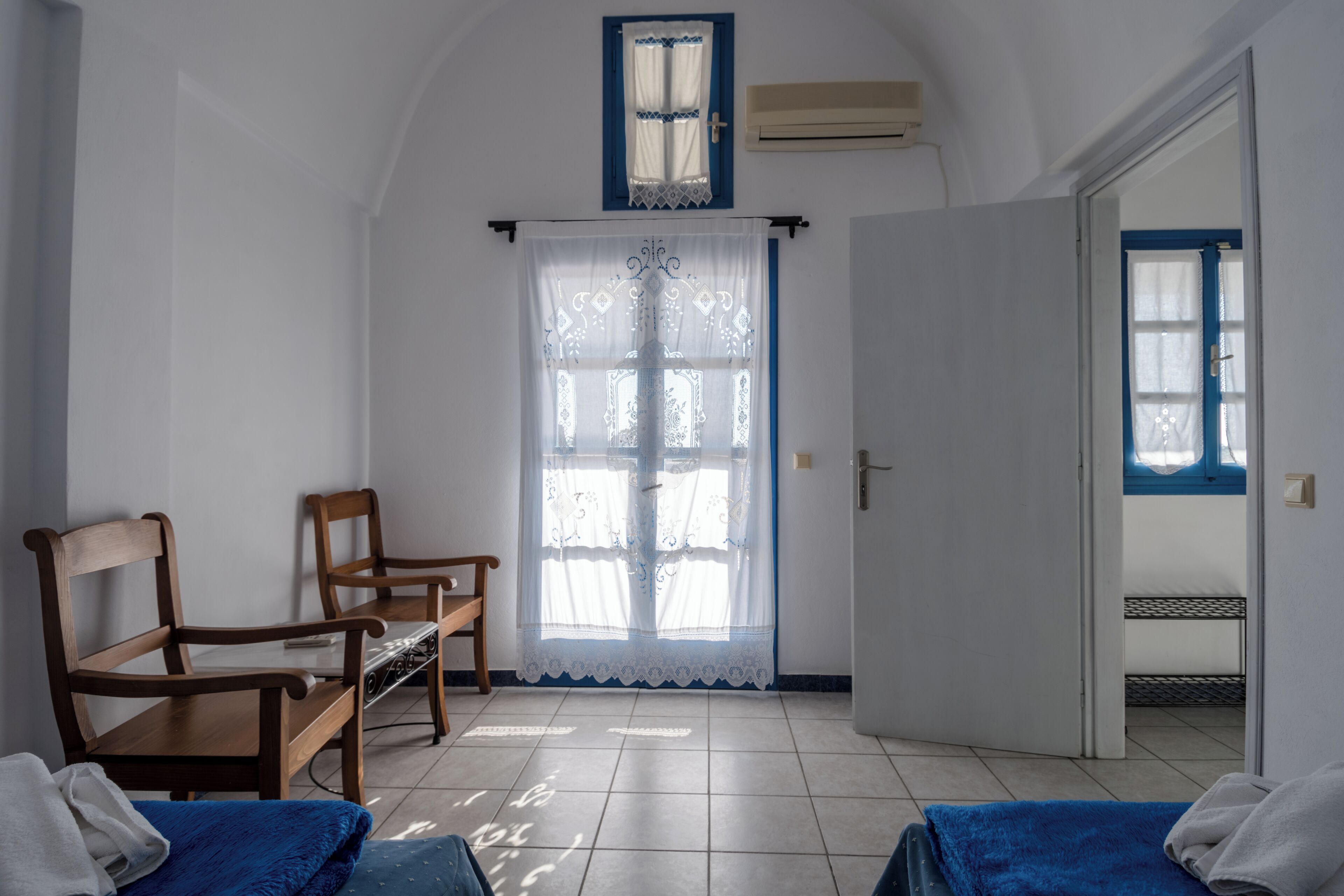 Een kamer met twee houten stoelen, een bed met een blauwe deken, een witte deur en een raam met een blauw kader.