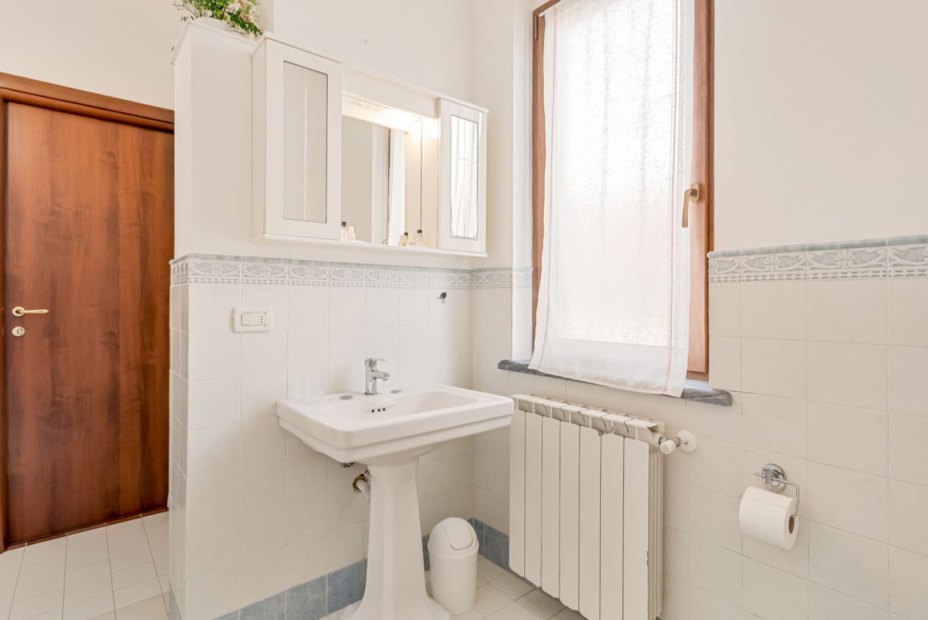 A bathroom with a pedestal sink, a wooden door, a window with curtains, a radiator, and a toilet paper holder.