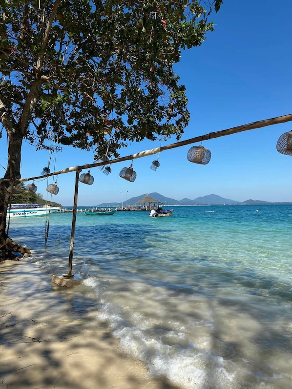 Uma praia com águas cristalinas, uma árvore e um barco ao fundo.