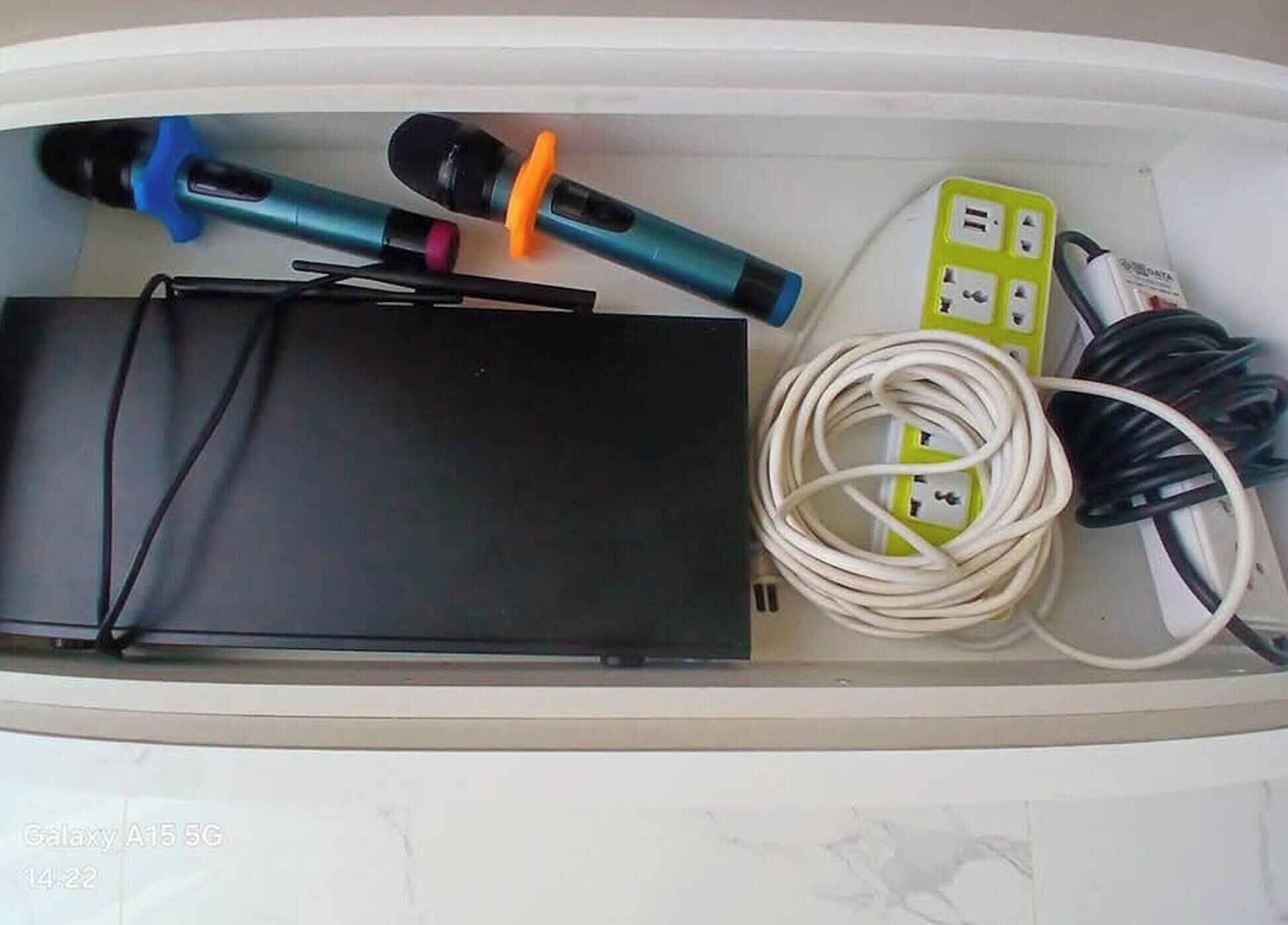 A drawer with a black electronic device, two blue and orange tools, a green power strip, and coiled white and black cables.