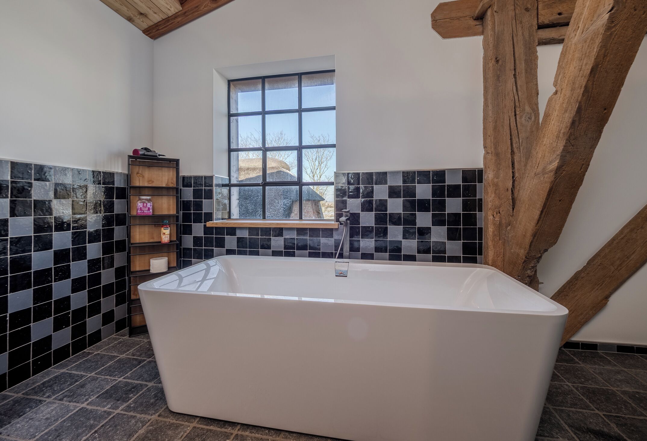 Un baño moderno con una bañera independiente, un estante de madera con artículos de aseo y una ventana que ofrece vistas al exterior.