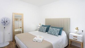 A neatly arranged hotel room with a bed, bedside table, lamp, and a standing fan. - Vv03. Atardecer Martianez (Puerto de la Cruz)