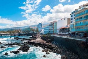 A coastal hotel with multiple floors, a rocky shoreline, and a clear sky. - Vv03. Atardecer Martianez (Puerto de la Cruz)