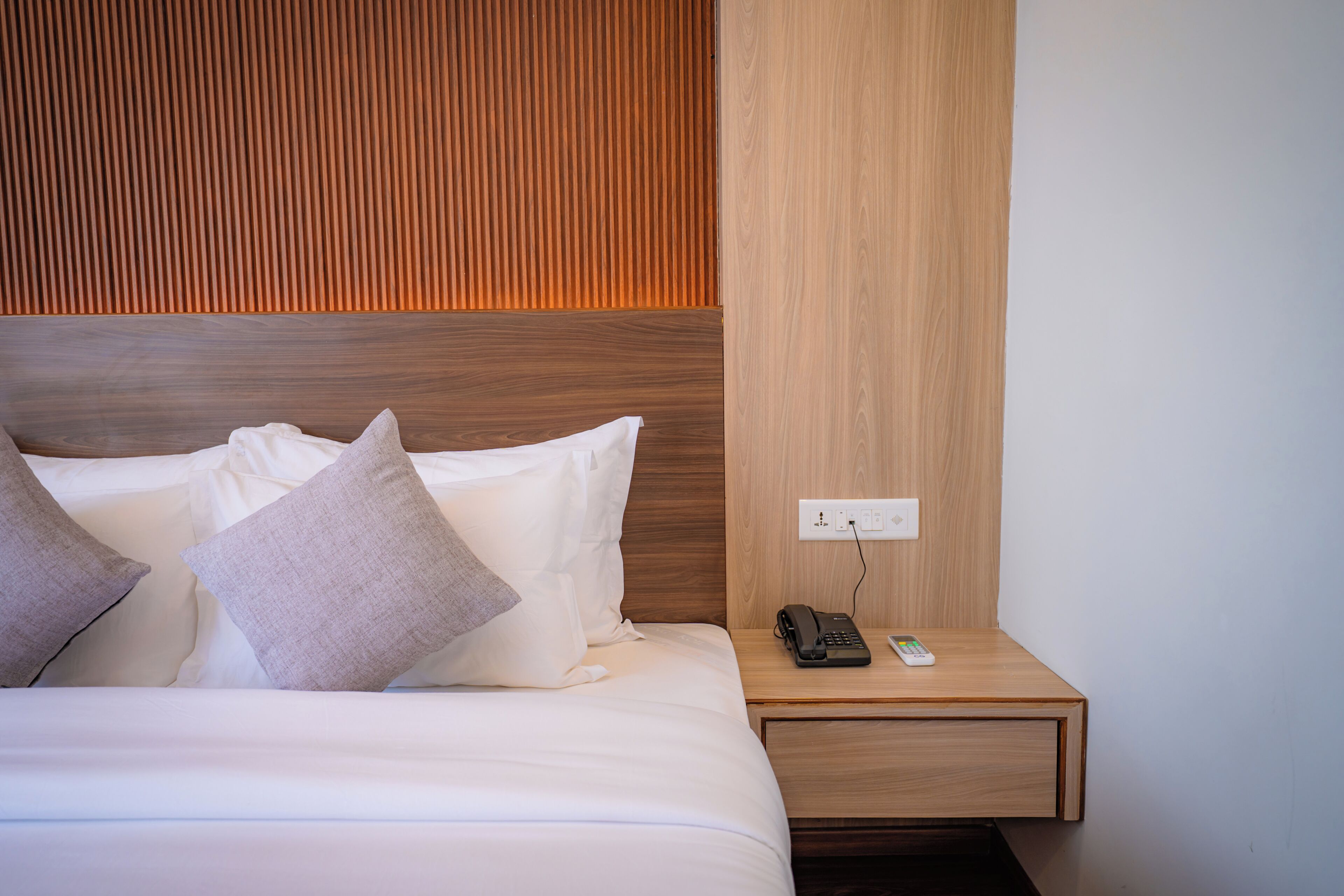 A neatly made bed with pillows, a bedside table with a phone and remote, and a wall with a wooden panel.