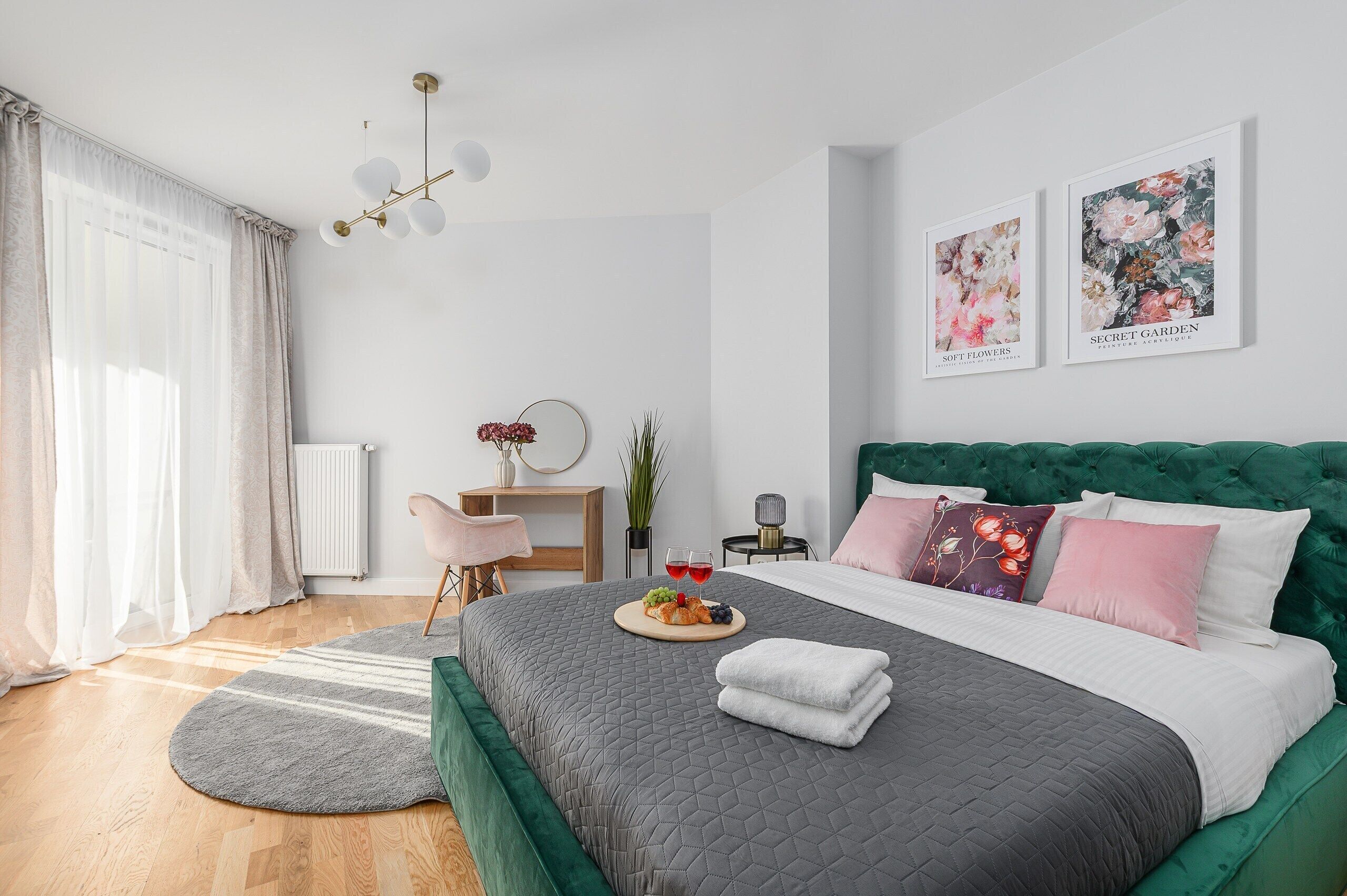 Ein Schlafzimmer mit Bett, Nachttisch, Sessel, Heizkörper, Fenster mit Vorhängen und zwei gerahmten Bildern an der Wand.
