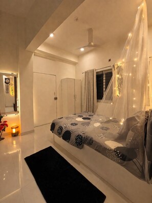 Une chambre à coucher avec un lit, un ventilateur de plafond, une fenêtre avec des rideaux et une table de chevet avec une lampe.