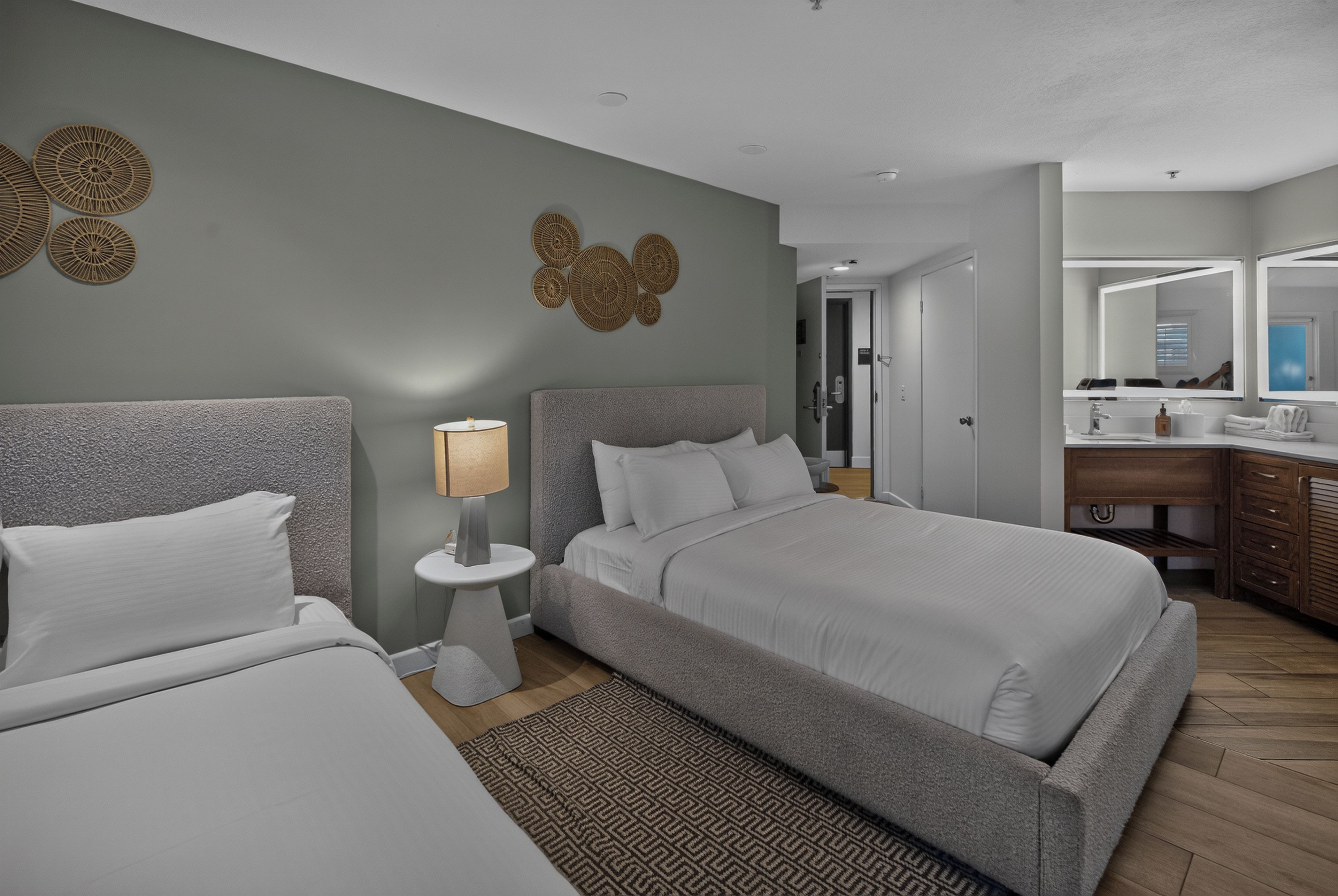 Een hotelkamer met twee bedden, een nachtkastje met een lamp, een badkamer met een spiegel en een kamerbreed tapijt met dessin.