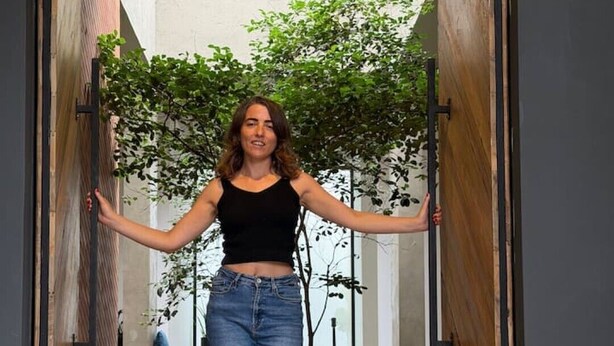 Uma pessoa em pé numa porta, com uma planta e um sofá ao fundo.