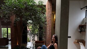 Uma mulher sentada em um banco em um interior moderno com uma árvore, uma mesa de madeira e uma cadeira branca.