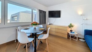 A modern living room with a dining area, a flat-screen TV mounted on the wall, a blue sofa, and a small side table with a plant. - Soley with parking by Interhome (De Haan)