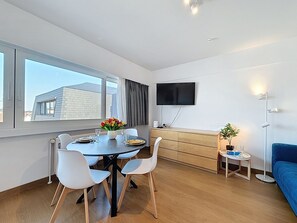 Un salon moderne comprenant un coin repas, un téléviseur à écran plat fixé au mur, un canapé bleu et une petite table d’appoint agrémentée d’une plante.