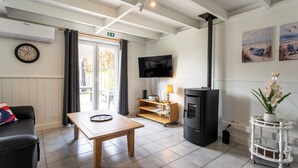 A room with a wooden table, a black sofa, a TV, a fireplace, and a window with curtains. - Alice Blue with airco & shared pool by Interhome (De Haan)