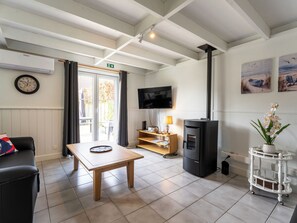A room with a wooden table, a black sofa, a TV, a fireplace, and a window with curtains. - Alice Blue with airco & shared pool by Interhome (De Haan)