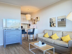 A modern living room with a sofa, coffee table, TV, and a kitchen area with a dining table and chairs. - Berolina 403 - Sand & Soul - Fantastic sunsets included! (Dahme)