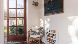 A room with a wooden door, a wicker chair, a white shelf with framed pictures, and a painting on the wall.