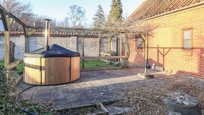 A wooden hot tub in a backyard with a brick house, a wooden pergola, and a garden area.