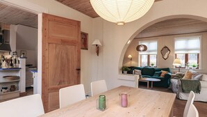 A spacious living area with a wooden dining table, a green sofa, and a kitchen visible through an open door.