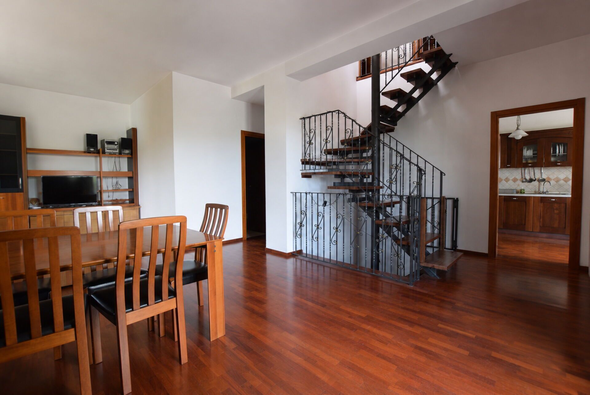 A spacious living area with a wooden dining table and chairs, a staircase with a wrought iron railing, and a kitchen area visible through an open doorway.
