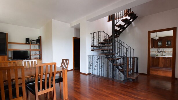Un vaste espace de vie comprenant une table et des chaises en bois, un escalier avec une rampe en fer forgé, et une cuisine ouverte sur le reste de la pièce.