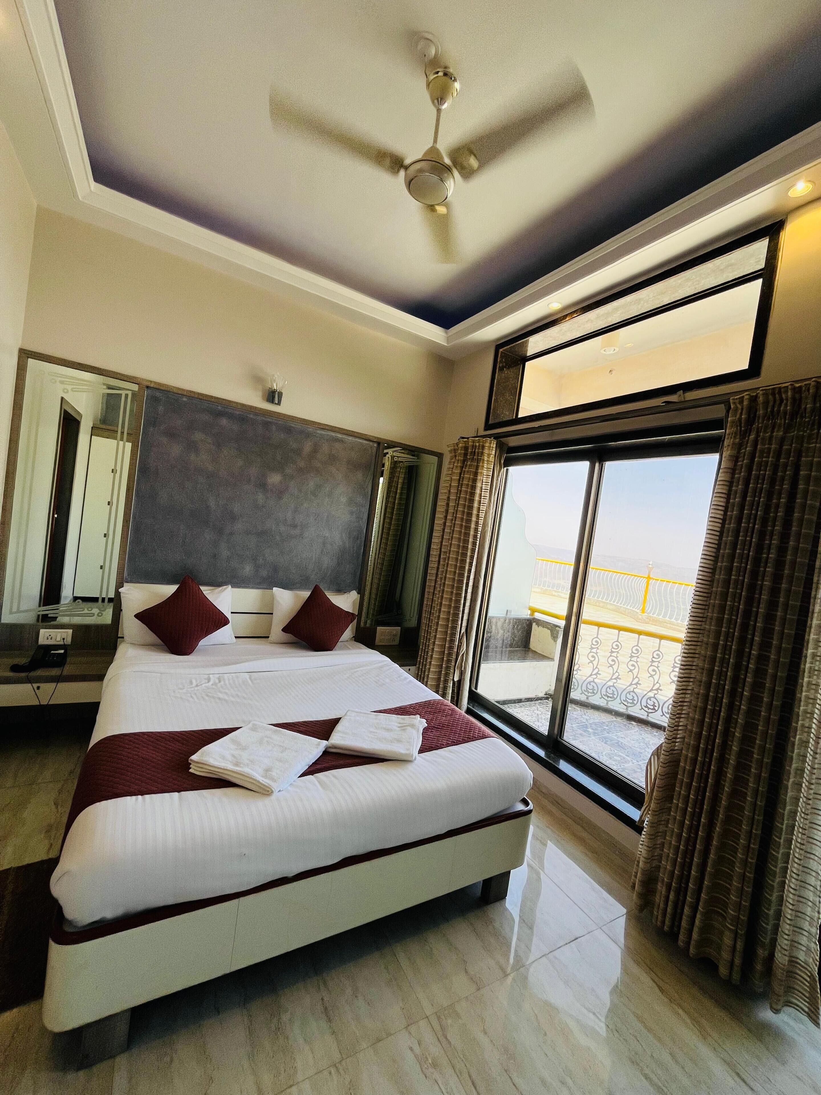 A hotel room with a large bed, a ceiling fan, a large window with curtains, and a balcony view.