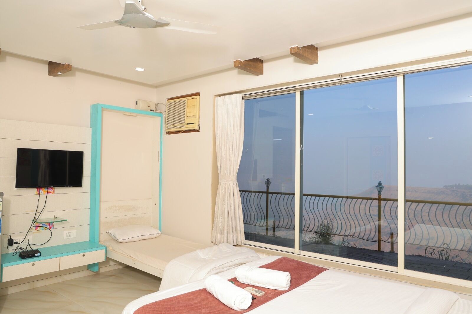 A hotel room with a bed, a TV, a window with a view, and a ceiling fan.