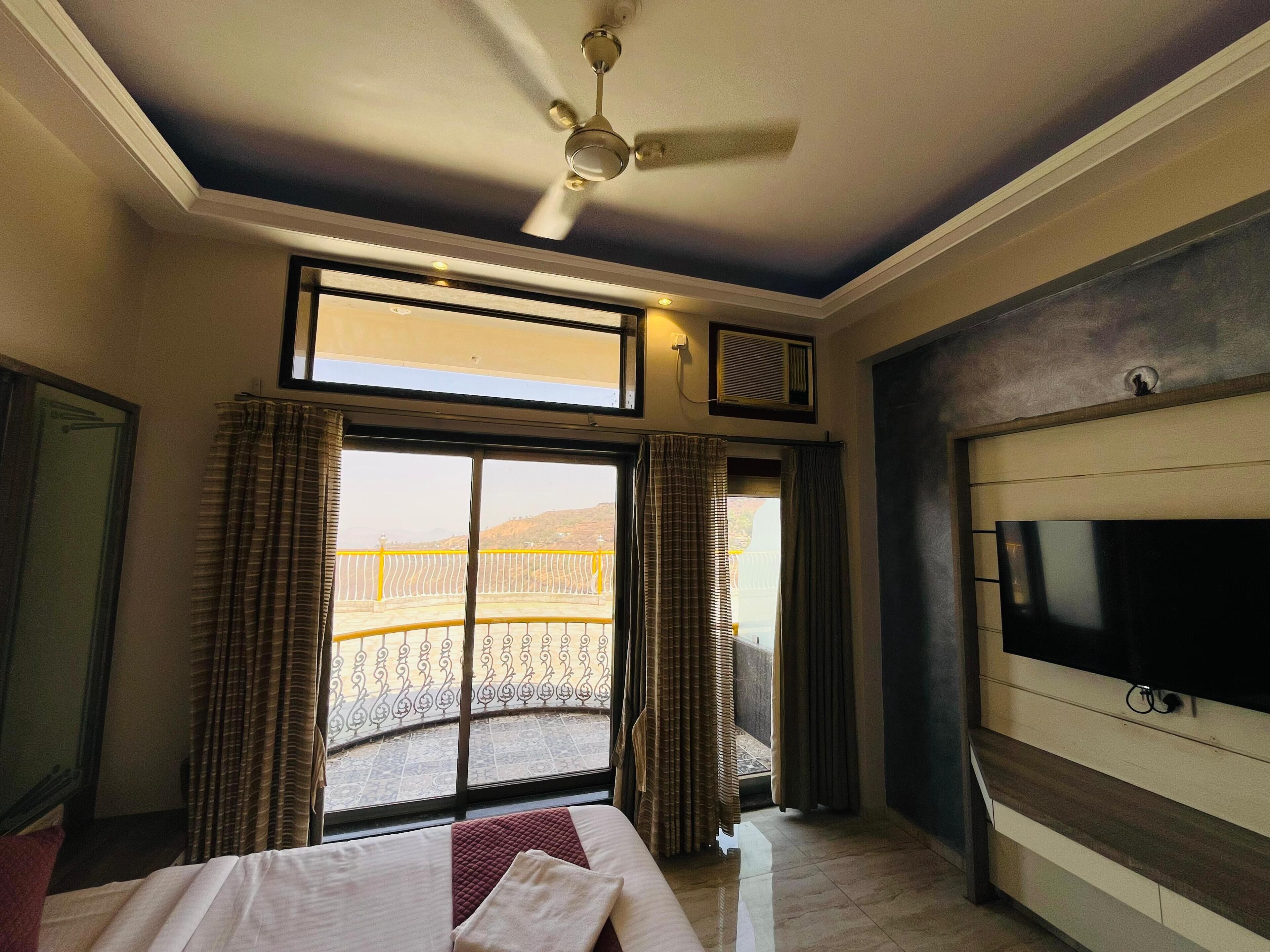 Habitación de hotel con cama, televisión, ventilador de techo, ventana con cortinas y vista a un balcón.