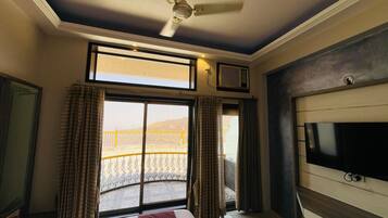 Habitación de hotel con cama, televisión, ventilador de techo, ventana con cortinas y vista a un balcón.