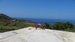 Paesaggio costiero con vista sul mare, una collina dalla vegetazione rada e una costruzione bianca con fiori rosa.