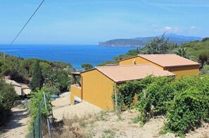 Un bâtiment jaune avec un toit en tuiles, entouré de verdure et offrant une vue dégagée sur la mer.
