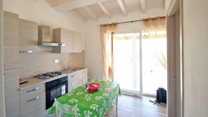 Une cuisine équipée d’une cuisinière à gaz, d’un four et d’un évier, une table à manger recouverte d’une nappe verte et une porte-fenêtre coulissante donnant sur l’extérieur.