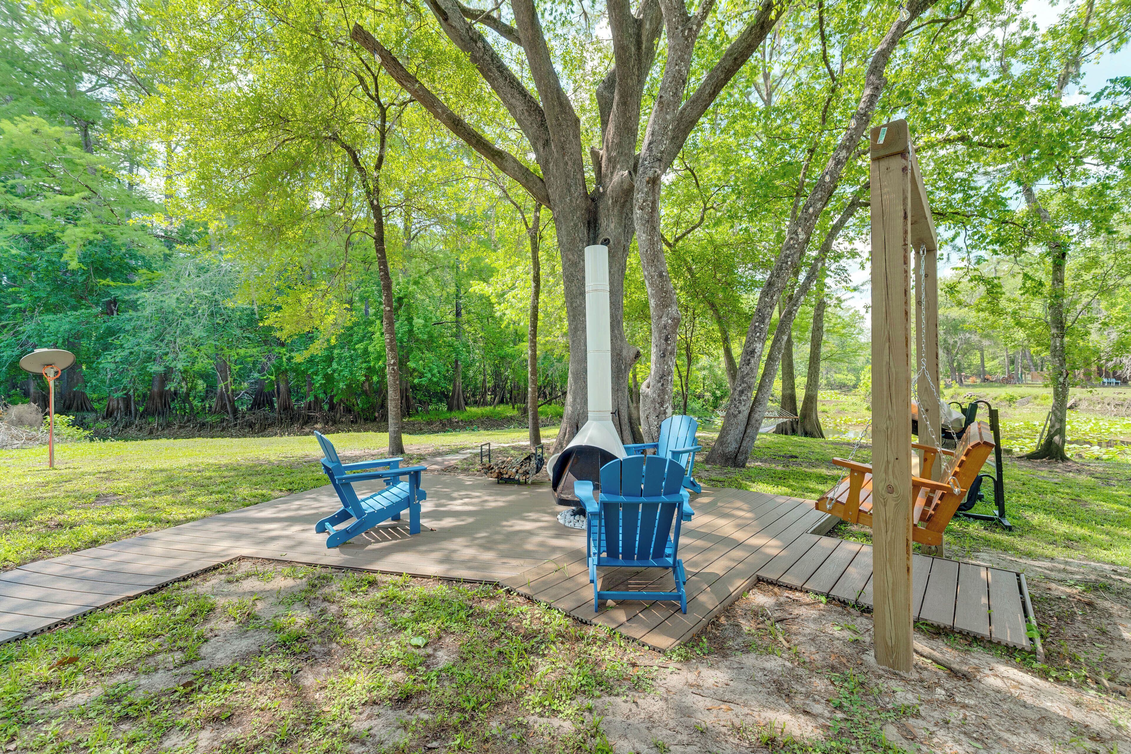 Una terraza de madera con sillas Adirondack, un brasero y un columpio en un área boscosa.