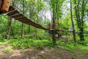 Un pont suspendu en bois, situé dans une zone boisée, avec une petite structure en bois en arrière-plan.