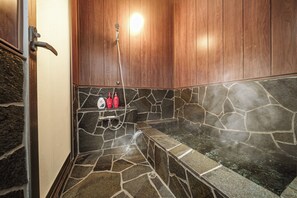 Uma sauna com piso de pedra, um banco, um chuveiro e garrafas vermelhas em uma prateleira.