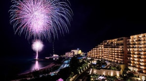Un hotel de varios pisos, vista a la playa y un espectáculo de fuegos artificiales en el cielo nocturno.