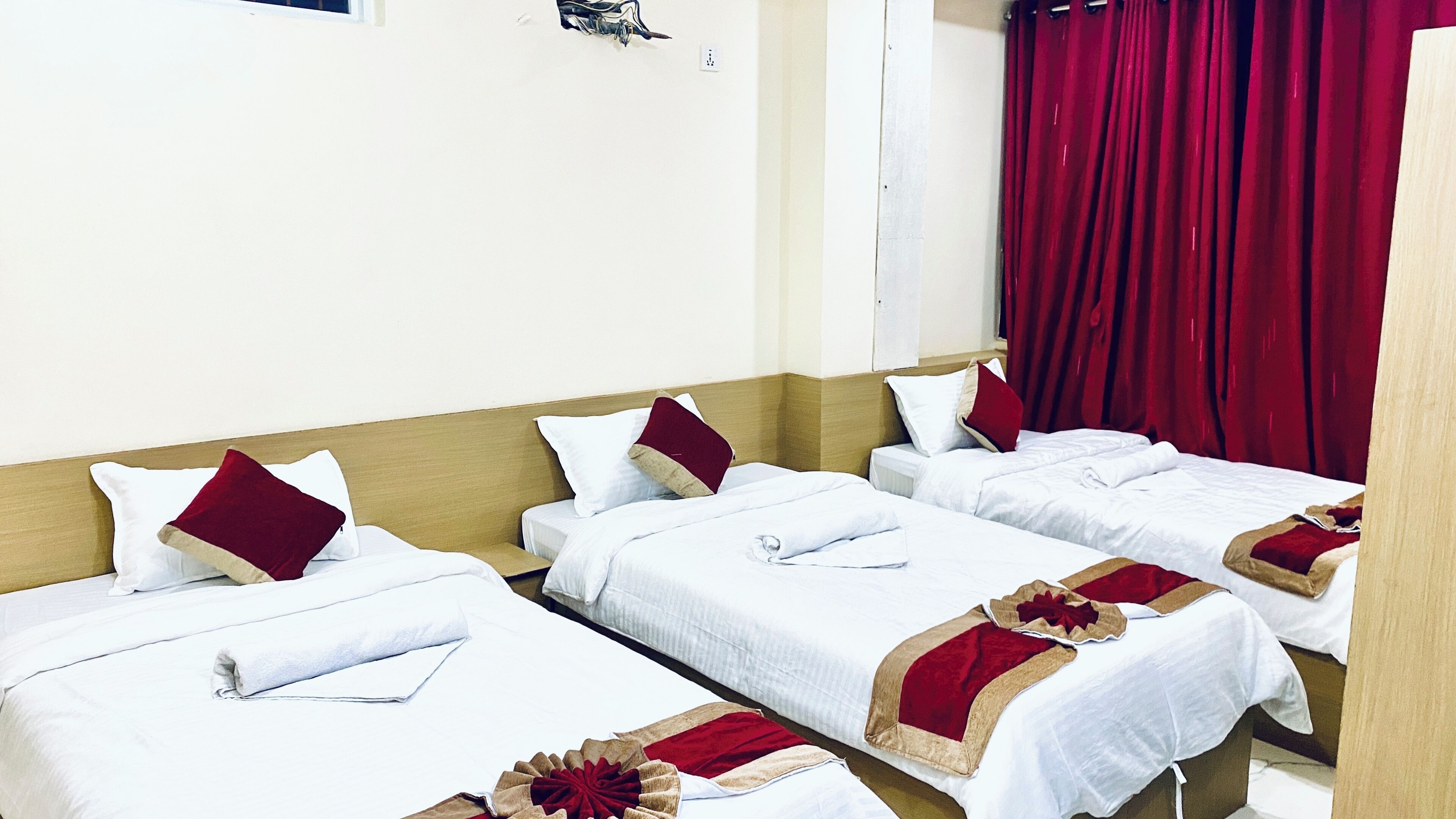 A hotel room with three single beds, each with white bedding and red accents, a wooden headboard, and red curtains.