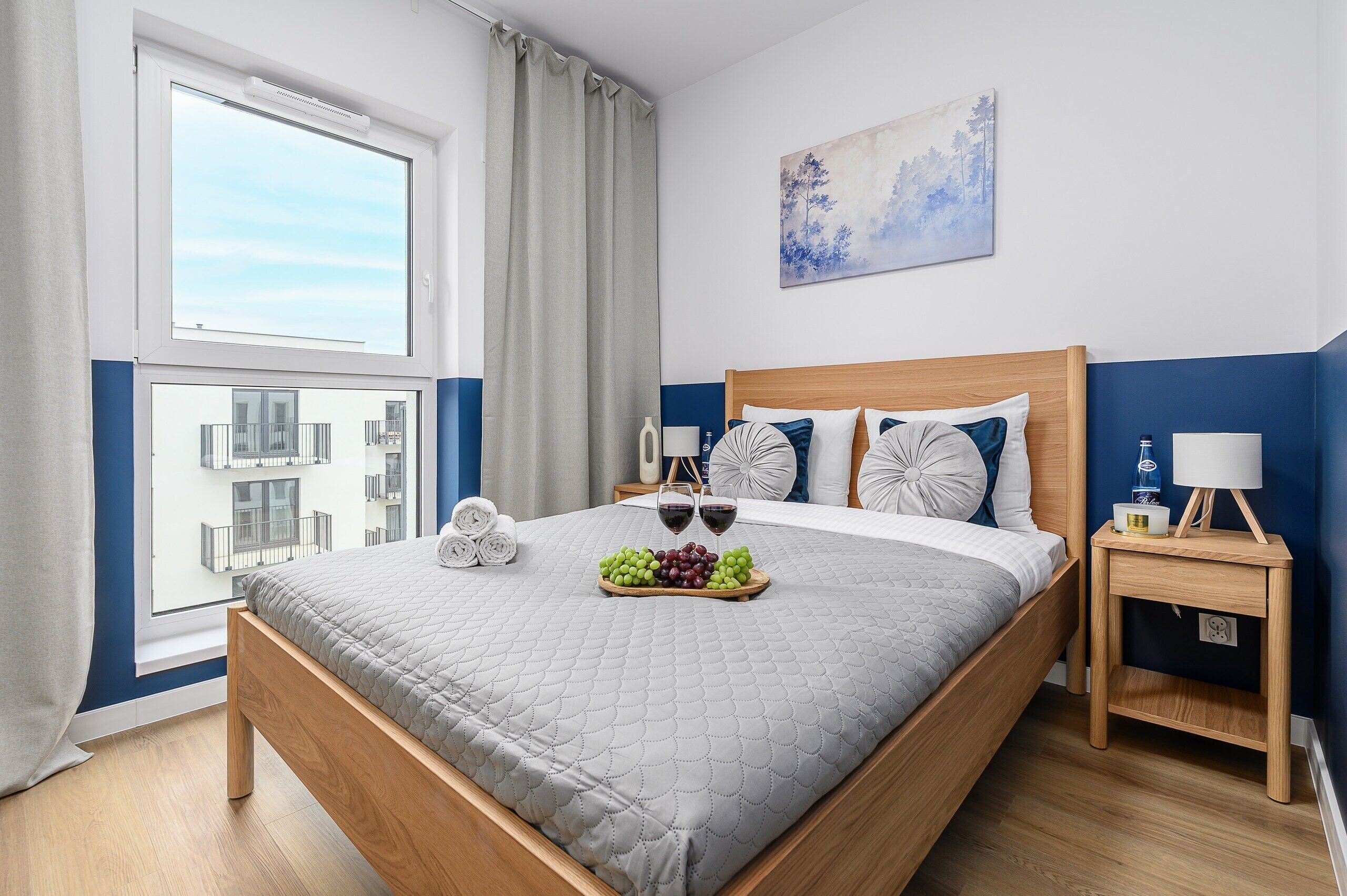 A neatly made bed with a quilted grey blanket, a bedside table with a lamp, and a fruit tray on the bed.