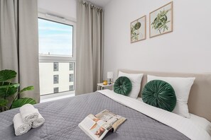 Une chambre d’hôtel avec un lit, une fenêtre avec des rideaux, une table de chevet avec une lampe, et deux reproductions encadrées de motifs botaniques accrochées au mur.