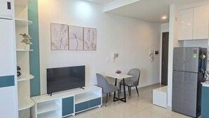 A modern living room with a flat-screen TV, a dining table with chairs, a refrigerator, and a wall-mounted shelf unit. - Way home studio sunrise city view (Thành phố Hồ Chí Minh)