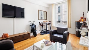 A modern living room with a flat-screen TV, a dining area with chairs, a scooter, a leather sofa, and a decorative cabinet. - 1BR Saint-Germain-des-Prés Seine Escape (Paris)