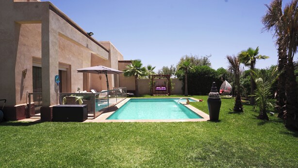 Une piscine avec un espace patio, un fauteuil suspendu et un petit jardin agrémenté de palmiers.