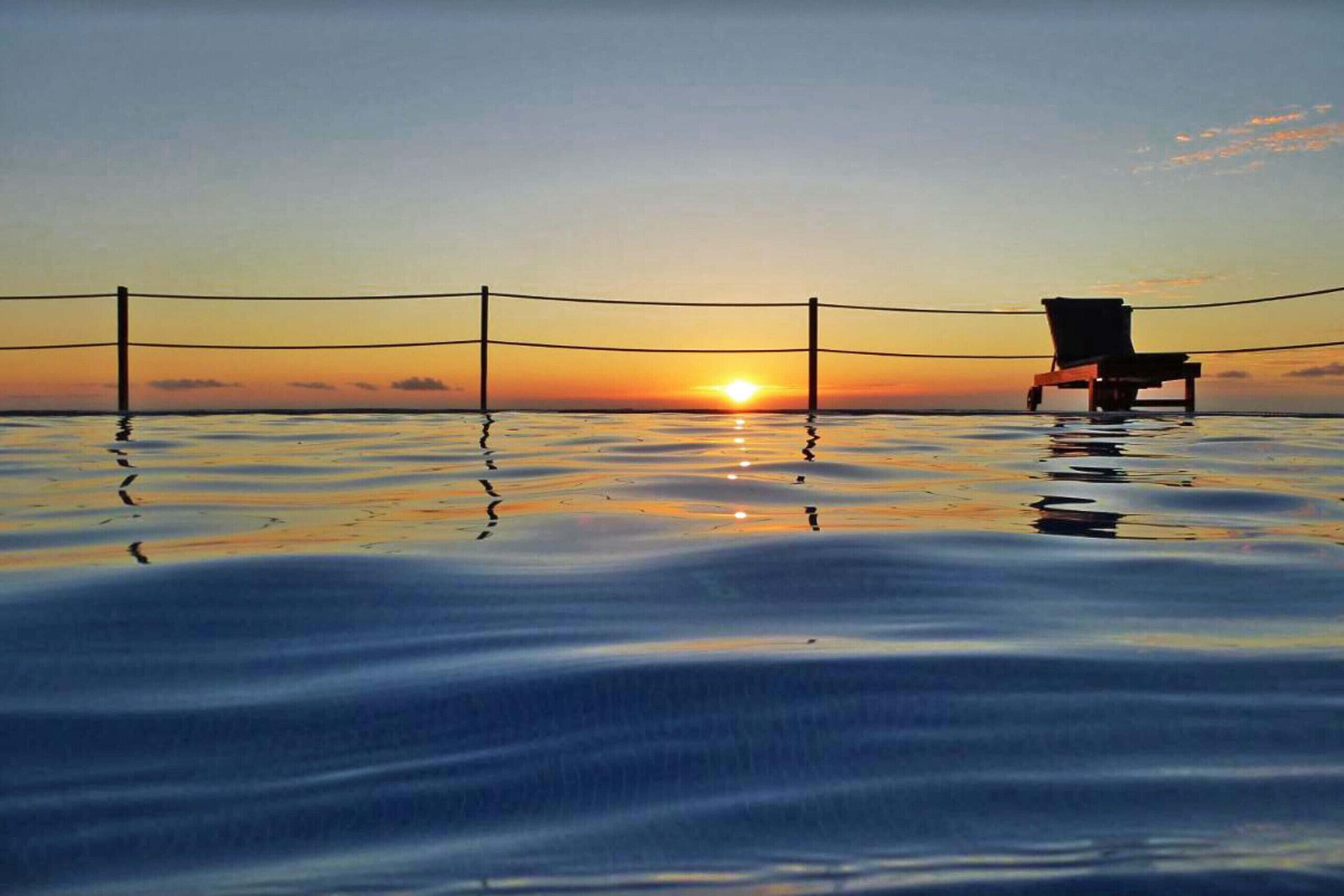 Ein Stuhl am Ufer des stillen Wassers bei Sonnenuntergang, im Hintergrund ein Zaun.