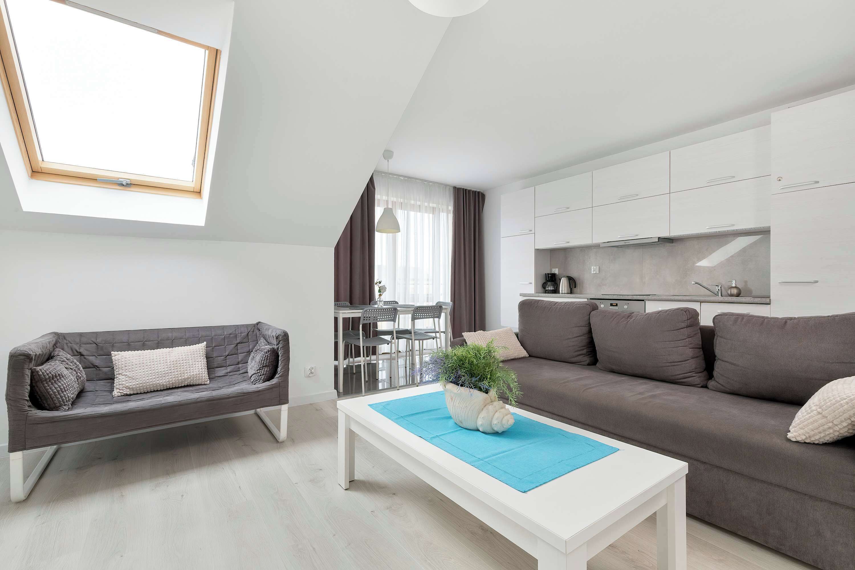 A bright living room features a dark grey corner sofa, a white coffee table with a blue runner, and a modern kitchenette in the background.