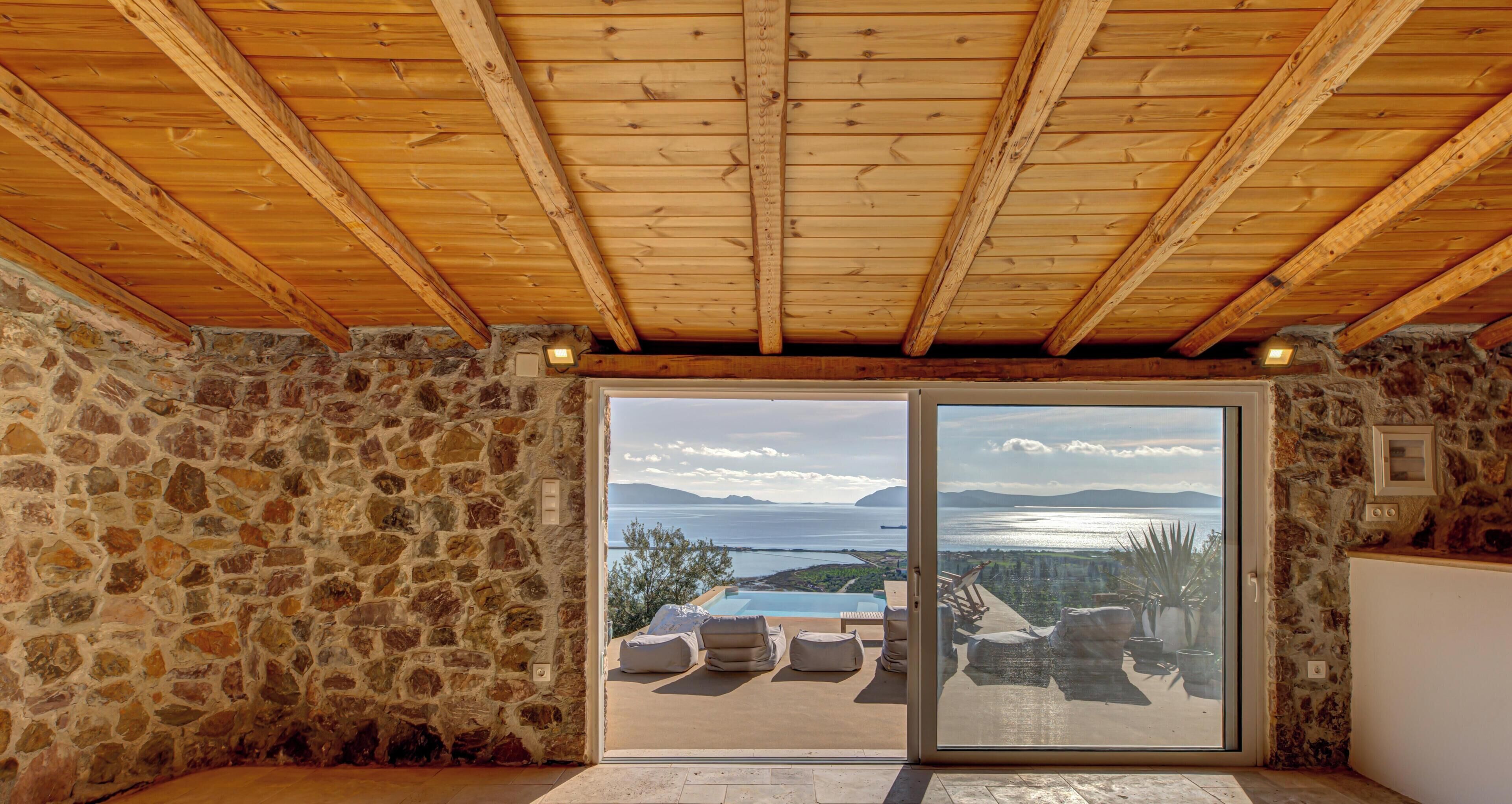 A stone-walled room with a wooden ceiling and a sliding glass door leading to a balcony with a pool and a view of the sea.