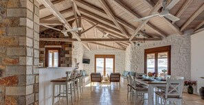 A rustic interior with exposed wooden beams, stone walls, a dining area with white furniture, and a view through a large window. - Villa Karima Sunset & Sea Views, Sea & Sunset View Villa Karima | Private Jacuzzi (Argolida)